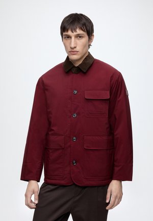 Jeune homme portant une veste bordeaux boutonnée avec plusieurs poches avant et un col marron foncé, debout devant un fond clair uni.