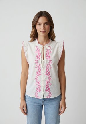 Femme portant un chemisier blanc sans manches avec une broderie florale rose à l'avant et un jean bleu clair, debout devant un fond uni.