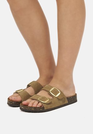 Pies con sandalias de ante marrón con dos grandes hebillas doradas y suela de corcho sobre un fondo claro y liso.