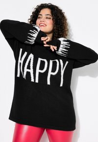Schwarzer Pullover mit weißem "HAPPY"-Text und gemusterten Bündchen, kombiniert mit glänzenden rosa Hosen. Hat ein lässiges, übergroßes Design.