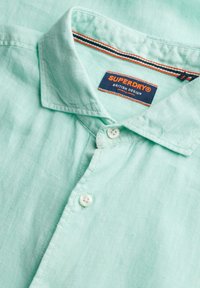 Camicia button-up verde menta chiaro con tessuto testurizzato. Presenta un colletto strutturato e un'etichetta con la scritta "SUPERDRY®". Due bottoni bianchi sono visibili.