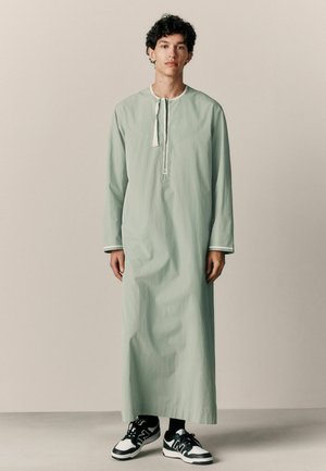 Tunica lunga verde menta con scollo rotondo e abbottonatura. Presenta maniche lunghe, spacchi laterali e rifinitura a contrasto. Indossata con sneakers nere.