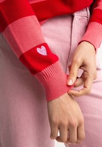 Červeno-růžový pruhovaný pletený svetr s žebrovanými manžetami, který má na rukávu malou vyšívanou srdíčko. V kombinaci se světle růžovými džíny.