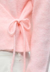 Pull rose doux et duveteux noué en nœud à la taille, porté avec un pantalon beige clair.