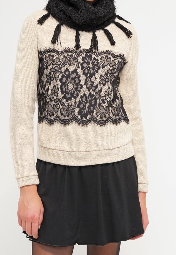 Pull beige avec superposition de dentelle noire et pompons, assorti à une jupe noire. Le pull pr ésente un tricot texturé, et la dentelle arbore des motifs floraux.