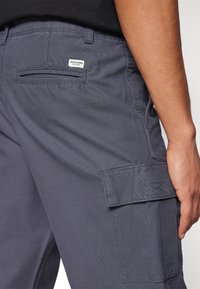 Pantalons cargo gris avec poches latérales et arrière, étiquette en tissu, et une texture durable et lisse. Des détails de couture sont visibles près des poches.