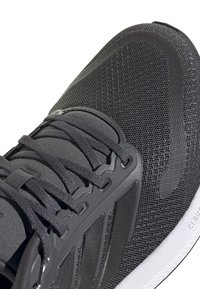 Gros plan d'une chaussure de sport noire avec une tige en mesh, des surcouches synthétiques texturées, des lacets noirs et un bord de semelle blanc coussiné.