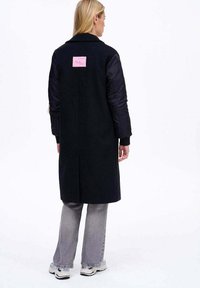 Žena otočená zády, oblečená v dlouhém černém kabátu s prošívanými rukávy, šedých džínách a bílých teniskách na bílém pozadí.