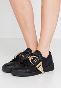 Baskets en cuir noir avec des accents dorés, comprenant un détail de boucle, un bout rond et une semelle texturée avec un logo le long du bord.