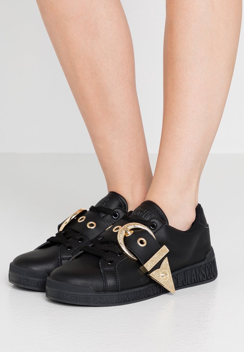 Baskets en cuir noir avec des accents dorés, comprenant un détail de boucle, un bout rond et une semelle texturée avec un logo le long du bord.