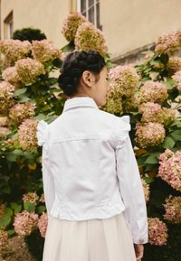 Nuori tyttö, jolla on letitetyt hiukset, pukeutuneena valkoiseen rypyttävään takkiin ja mekkoon, seisoo kukkivien vaaleanpunaisten hortensiapensaineiden edessä ulkona.
