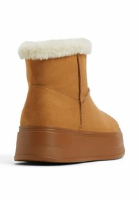 Botas de tobillo de ante marrón con un cuello de felpa blanca, que presentan una gruesa suela de plataforma marrón y una textura suave. Detalles de costura reforzada presentes.