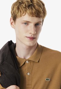 Camisa polo marrón con una tela texturizada, tres botones y un pequeño logo de cocodrilo verde en el pecho. El modelo sostiene una chaqueta oscura.