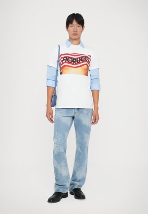 Young man wearing white graphic T-shirt over blue shirt, faded light blue jeans with patterns, black shoes, and carrying blue shoulder bag.