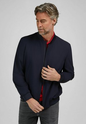 Man with gray hair fastening zipper on dark navy bomber jacket, wearing red shirt and gray pants, standing against light gray background.