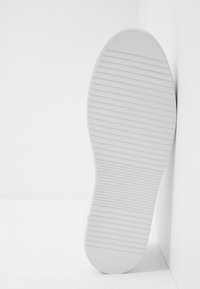 Bílá podrážka tenisky s texturovaným, vroubkovaným vzorem a zaoblenou špičkou. Obuv je umístěna na bílém pozadí.