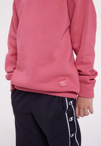 Persona che indossa un felpa rosa con l'etichetta "Sii il tuo campione" e pantaloni della tuta neri con strisce bianche del logo Champion sui lati.