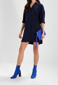 Donna che indossa un vestito camicia blu navy con maniche arrotolate, mentre tiene una pochette blu e indossa stivaletti blu brillante con tacco a blocco.