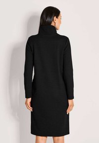 Frau mit langen dunklen Haaren, die ein schwarzes langärmeliges Rollkragenkleid trägt und mit dem Rücken zu einem schlichten hellen Hintergrund steht.
