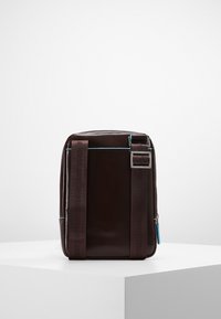 Borsa in pelle marrone scuro di forma rettangolare, con spallacci in tessuto regolabili, minuteria metallica e un dettaglio della cerniera blu a contrasto.