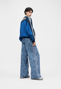 Personne portant un sweat-shirt Adidas bleu et un jean à jambes larges avec motif cachemire, debout en angle avec une main dans la poche sur fond blanc.