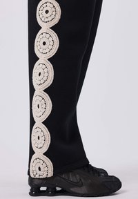 Schwarze Jogginghose mit einem cremefarbenen Häkelbesatz und kreisförmigen Mustern an der Außenseite, kombiniert mit schwarzen Sportschuhen.