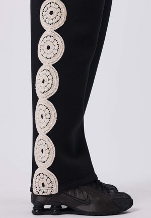 Zwarte trainingsbroek met een crèmekleurige haakjesafwerking met cirkelvormige patronen aan de buitenkant, gecombineerd met zwarte sportschoenen.
