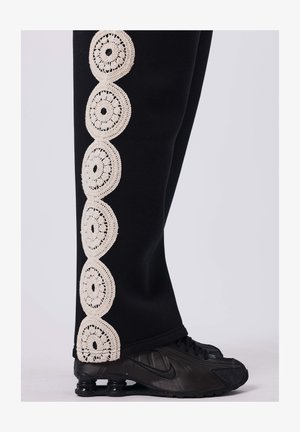 Zwarte trainingsbroek met een crèmekleurige haakjesafwerking met cirkelvormige patronen aan de buitenkant, gecombineerd met zwarte sportschoenen.