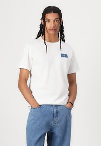 Vit bomulls-t-shirt med en blå grafisk logo på vänster sida, tillsammans med ljusblå denimjeans. Avslappnad passform och korta ärmar.