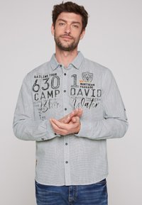 Camisa de botones de color gris claro con un patrón texturizado, que presenta gráficos impresos en negro y naranja, y botones de contraste a lo largo del trazo.