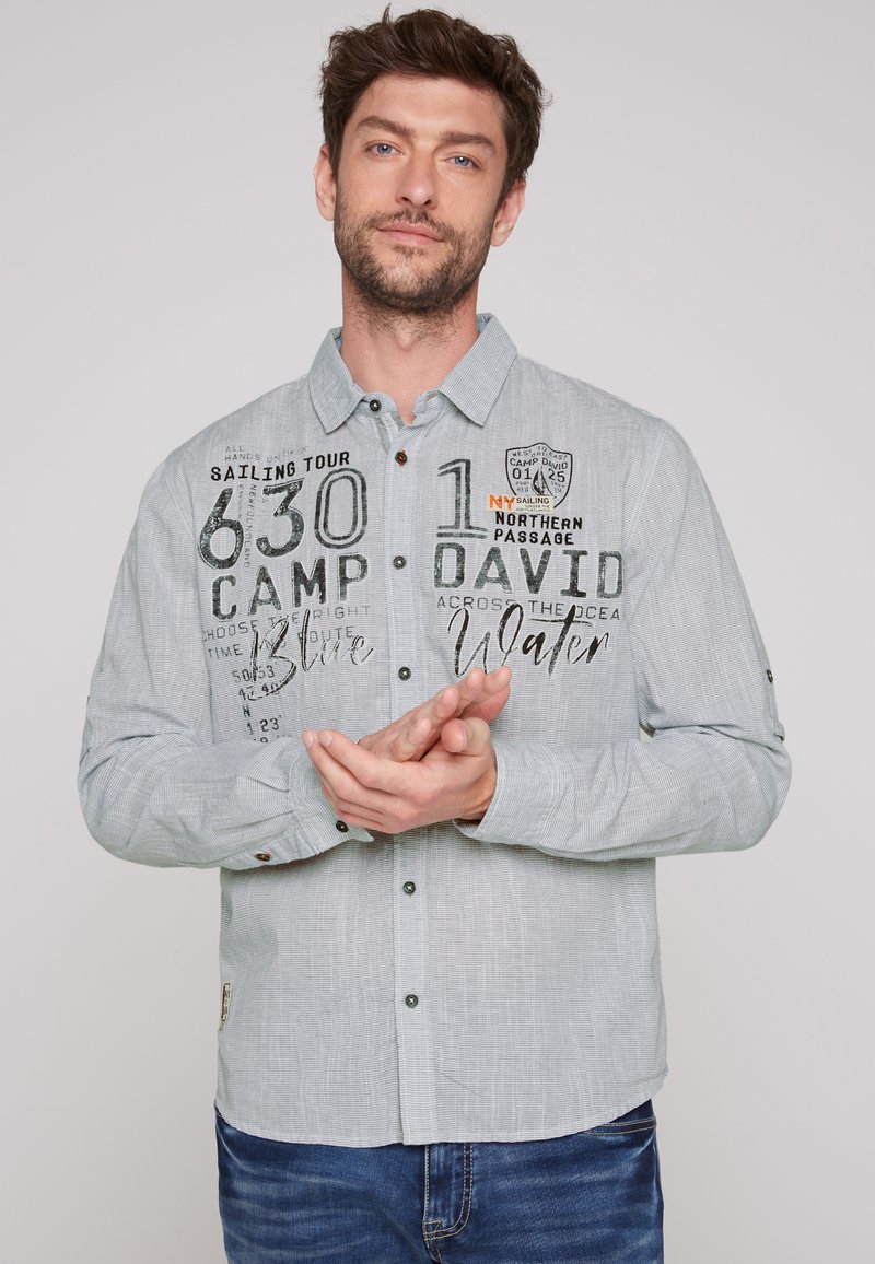 Camisa de botones de color gris claro con un patrón texturizado, que presenta gráficos impresos en negro y naranja, y botones de contraste a lo largo del trazo.