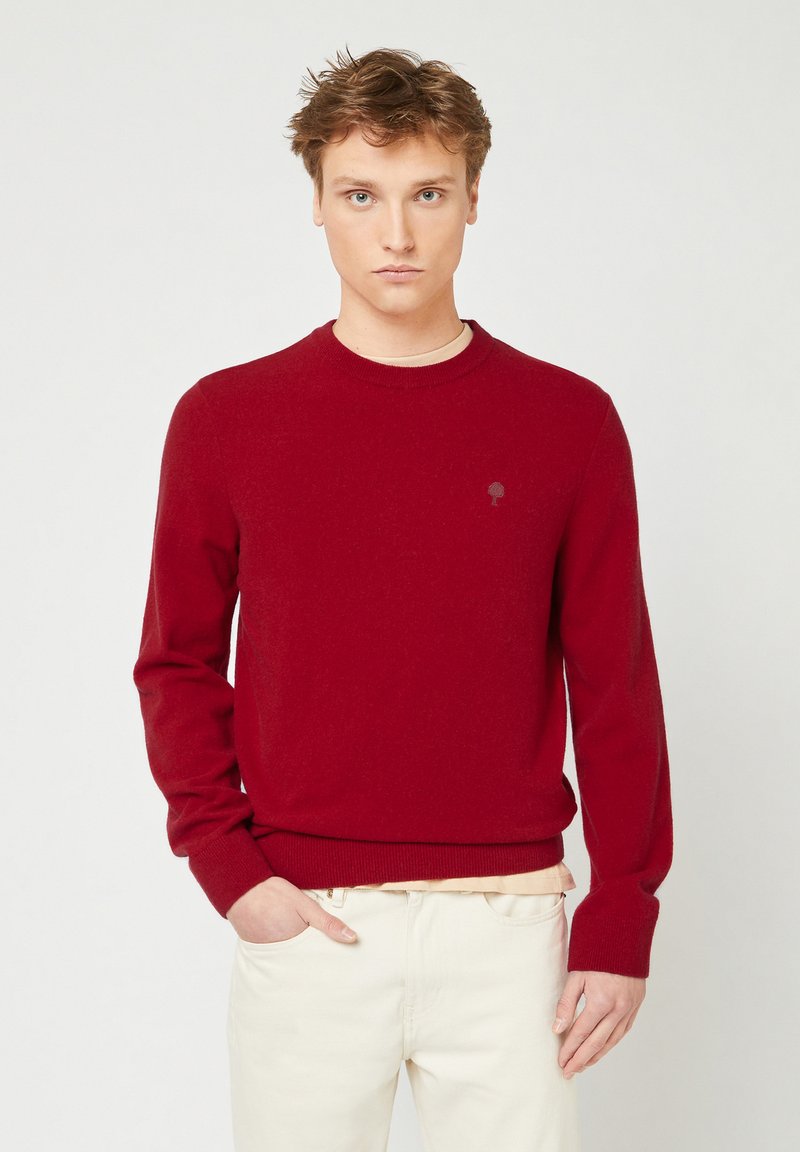 Maglione rosso a maglia con scollo rotondo e maniche lunghe, caratterizzato da un sottile logo ad albero e polsini e orlo a costine. Indossato con pantaloni chiari.