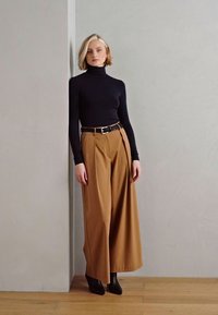 Top negro de cuello alto, pantalones anchos color tostado con pliegues, cinturón negro y botas de tacón negras sobre un fondo neutro. Texturas suaves.