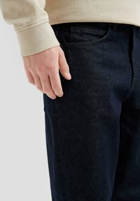 Tumed sinine teksapüksid, millel on keha lähedane lõige, koos heleda beeži pikkade varrukatega särgiga. Näha on taskud ja tekstuurilised kangaste detailid.