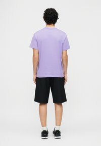 Άτομο με σγουρά σκούρα μαλλιά στέκεται με την πλάτη στραμμένη φορώντας λιλά κοντομάνικο μπλουζάκι, μαύρο σορτς, άσπρες κάλτσες και μαύρα αθλητικά παπούτσια.