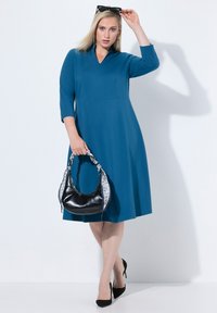 Turkusowa sukienka do kolan z rękawami 3/4 i dekoltem w szpic. Połączona z czarną torebką na ramię z białym wzorzystym paskiem i czarnymi szpilkami.