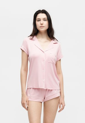 Νεαρή γυναίκα με ροζ ανοιχτό κοντομάνικο πουκάμισο πιτζάμα με κουμπιά και ταιριαστό σορτς, στέκεται μπροστά σε απλό λευκό φόντο.
