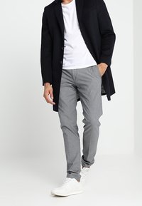 Schwarzer Mantel über einem weißen T-Shirt; graue gemusterte Hose; weiße Sneaker. Verfügt über eine maßgeschneiderte Passform und geschichtete Texturen.