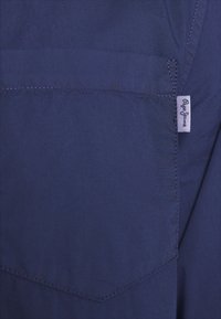 Nærbillede af en mørkeblå stoflomme med detaljeret syning og et lille hvidt stofmærketag på højre kant.