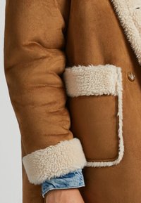 Brun kappa med en mjuk, mockaliknande textur. Den har en fluffig, krämfärgad krage och manschetter, samt en lapplåda på sidan.