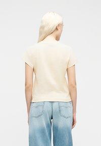 Γυναίκα με μακριά ξανθά μαλλιά που φοράει μπεζ μπλούζα με κοντά μανίκια και ανοιχτό μπλε τζιν παντελόνι, γυρισμένη προς τα πίσω με φόντο έναν απλό τοίχο.