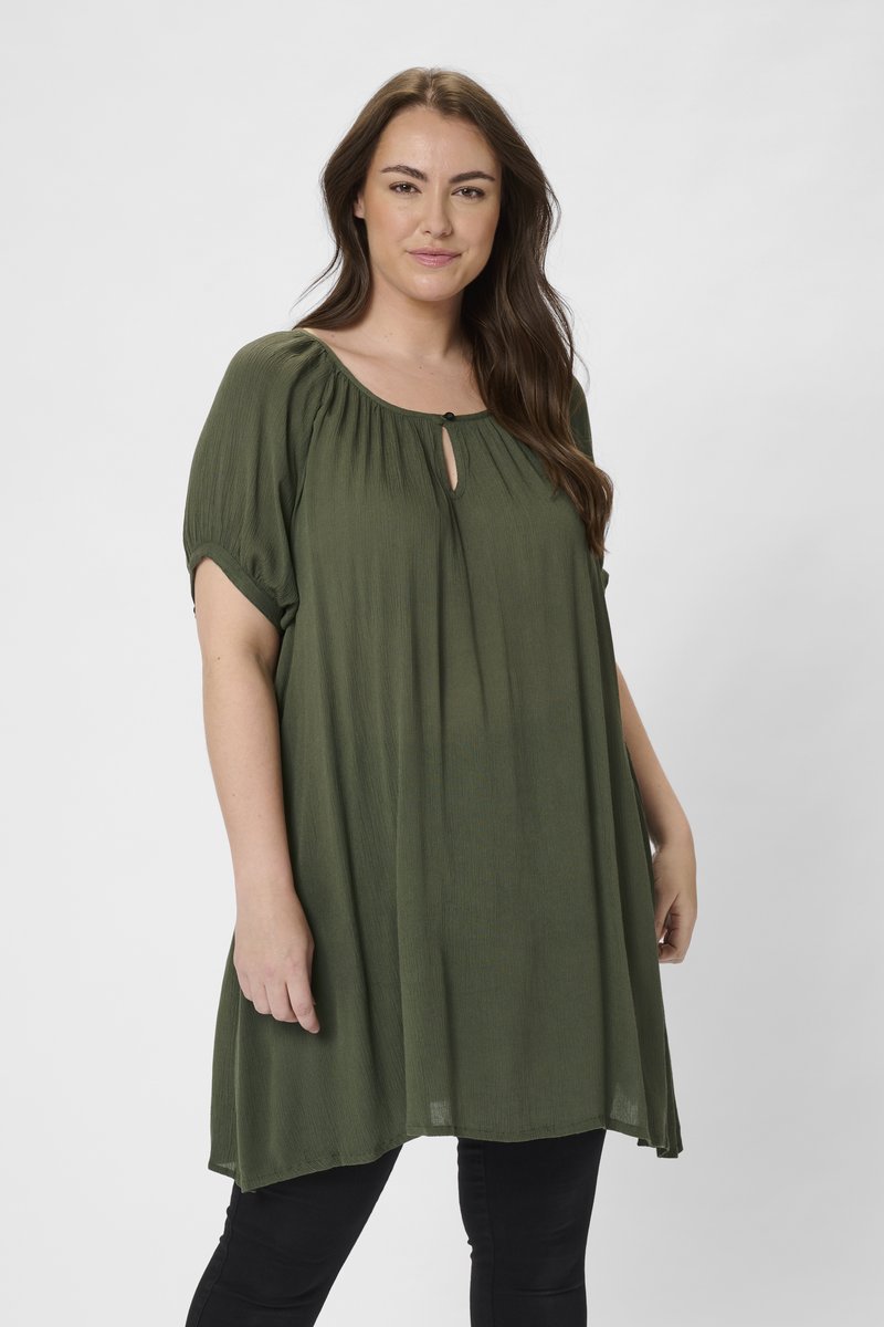 Tunique verte surdimensionnée avec un décolleté en trou de serrure et un buste froncé. Fabriquée dans un tissu léger avec des manches courtes. Associée à un pantalon noir.