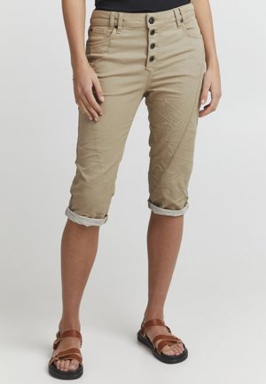 Pantalons cropped beiges en tissu texturé avec une fermeture à boutons et des poignets retroussés, associés à des sandales brunes à lanières.