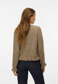 Brūns adīts džemperis ar ribām piedurknēs un volāna apakšu, ar vienkāršu tekstūru. Valkāts kopā ar tumšiem džinsa džinsiem. Skats no aizmugures.