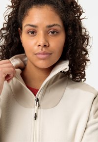 Woman with curly hair wearing a beige fleece jacket, holding the collar with her left hand against a plain white background.