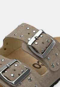 Μπεζ σουέντ σανδάλι με δύο ρυθμιζόμενες ασημί αγκράφες, διακοσμητικά ασημί καρφιά και επενδεδυμένη σόλα με φυσικό φινίρισμα.