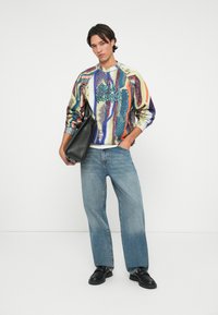 Strikket sweater med multicolor vertikale mønstre og grafisk tekst, parret med lyseblå bredbenede jeans og sorte lædersko, holder en sort taske.