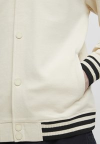 Cream-colored sweatshirt with a smooth texture, featuring a button front and black-striped ribbed cuffs. Includes side pockets.