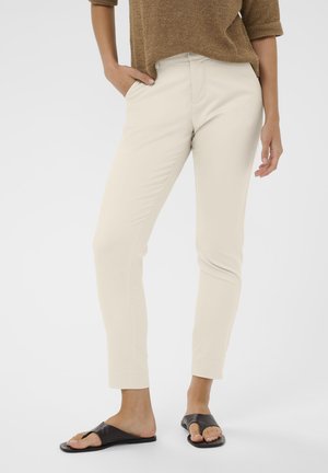 Kvinde iført beige figursyet bukser, brun striktop og sorte åbne flade sandaler med én hånd i lommen mod hvid baggrund.