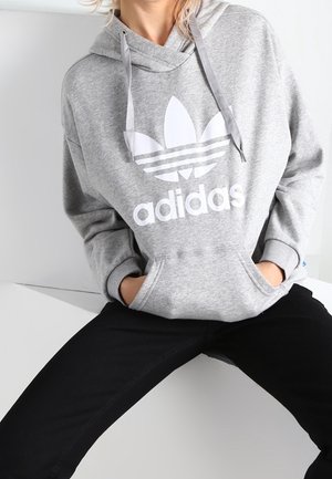 Osoba ubrana w szarą bluzę Adidas z kapturem z białym logo i czarne spodnie, siedząca z rękami w kieszeni bluzy na białym tle.